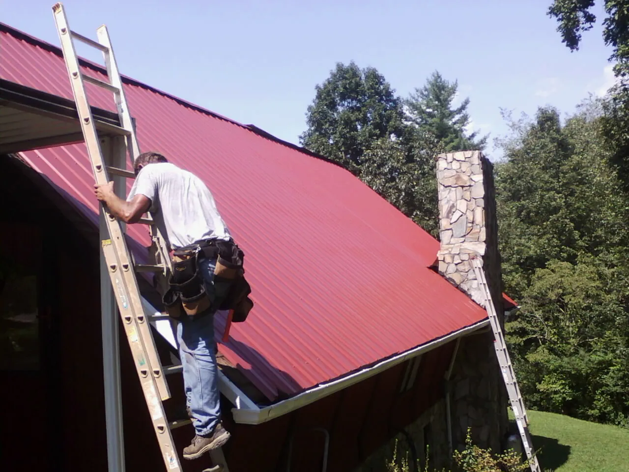 Roofer on a ladder beside a red metal roof and stone chimney — Soffit & Fascia Repair by Roofing Brothers in Harsens Island